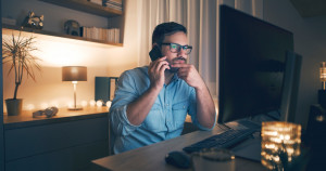 Foto di un uomo preoccupato che usa il suo cellulare, mano sul viso, guardando il monitor del suo computer