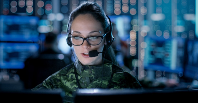 A woman seated in a dimly lit room full of monitors wearing a headset with a microphone by her mouth