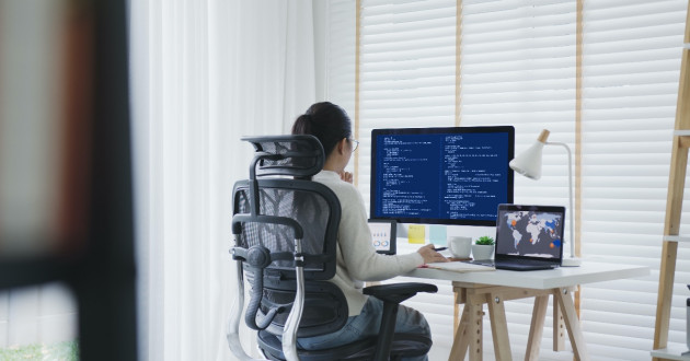 A woman with her back to us seated in a high-back chair at a home office desk working on a computer