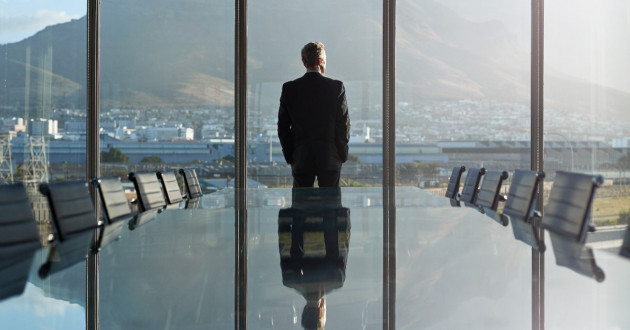 A male CEO standing at the edge of a large conference table with his back to us looking out of wall-size windows