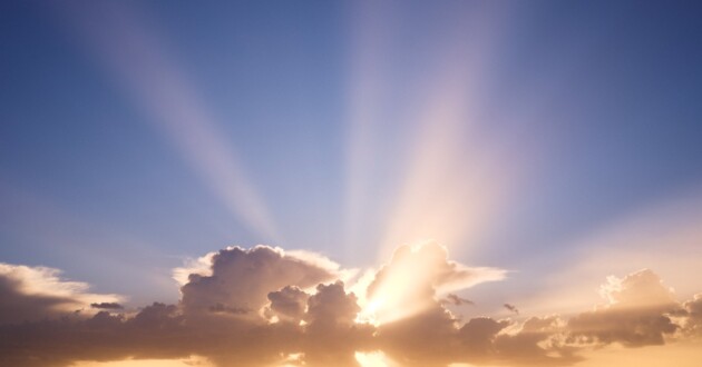 White fluffy clouds in a blue sky with the sun and rays coming through the clouds