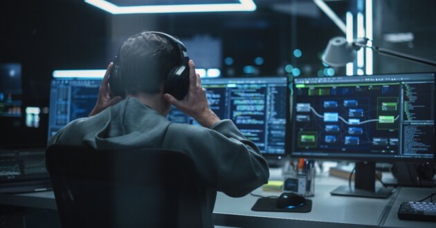 A man sitting at a desk with multiple monitors displaying audio waves wearing headphones with his back to us