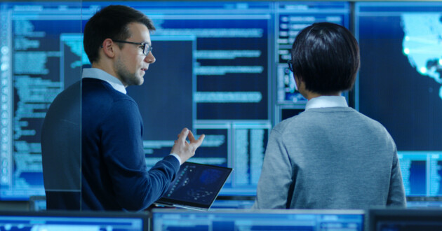 Two IT techs discussing the ONCD Report On The Cybersecurity Posture Of The U.S. in front of a large blue display.