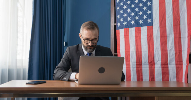 A caucasian businessman in a suit sitting at a desk in front of an American flag working on a laptop