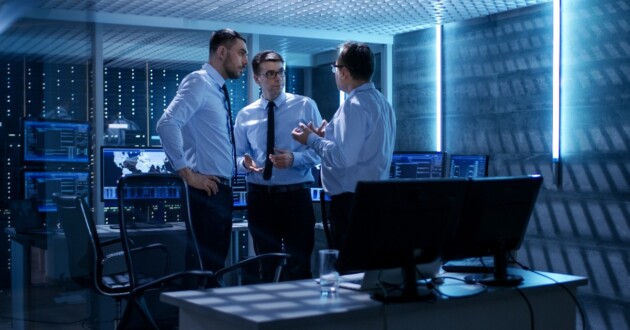 3 businessmen standing in a dimly blue-lit office surrounded by computers talking