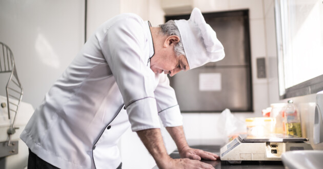 A senior man in a chef uniform standing hunched over in a commercial kitchen