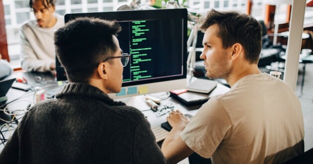 rear view of 2 male programmers at a desk discussing coding on a computer